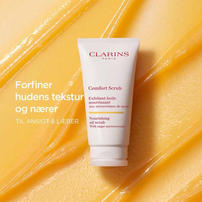 Clarins Comfort Scrub til ansigt og l&aelig;ber p&aring; en orange, tekstureret baggrund, der fremh&aelig;ver dens forfinende og n&aelig;rende effekt.