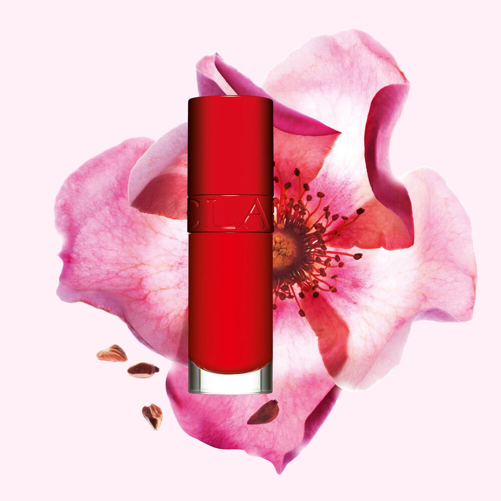 Packshot af Clarins Lip Comfort Oil i Iconic Red, placeret ved siden af en hybenroseblomst p&aring; en sort-r&oslash;d baggrund.