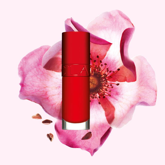 Packshot af Clarins Lip Comfort Oil i Iconic Red, placeret ved siden af en hybenroseblomst p&aring; en sort-r&oslash;d baggrund.