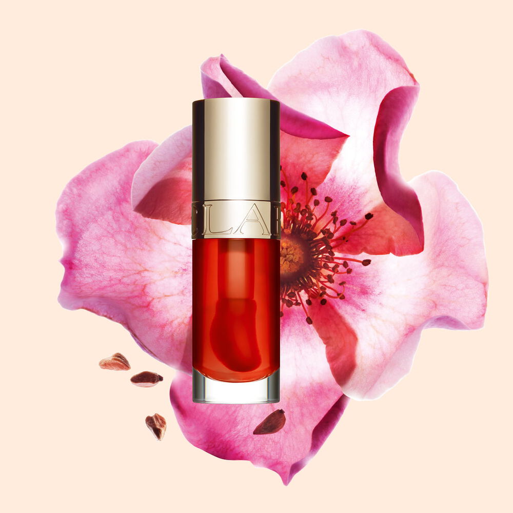 Packshot af Clarins Lip Comfort Oil i Apricot, placeret ved siden af en nyponrose p&aring; en lys orange ferskenfarvet baggrund.