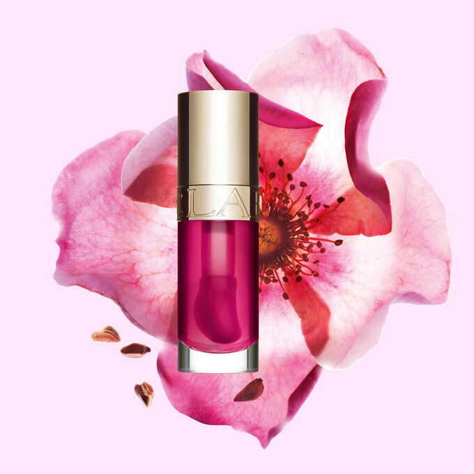 Packshot af Clarins Lip Comfort Oil i Raspberry, placeret ved siden af en nyperose p&aring; en lys ferskenfarvet baggrund.