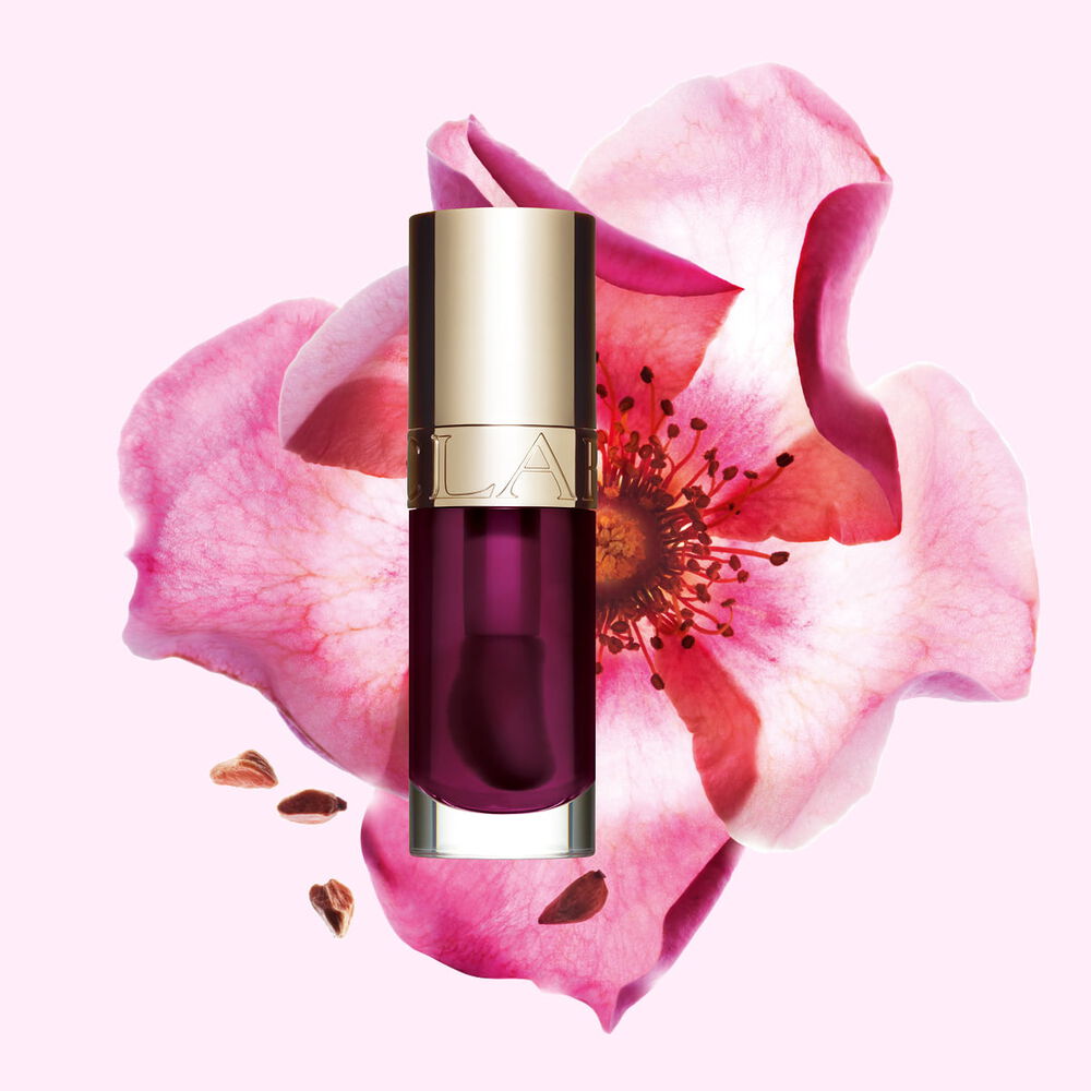 Packshot af Clarins Lip Comfort Oil i Plum, placeret ved siden af en nyrose p&aring; en lys rosa baggrund.